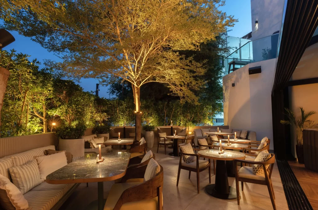 HAUS (4) garden terrace seating with marble tables, lush uplighted trees, and neutral-toned lounge sofas at night